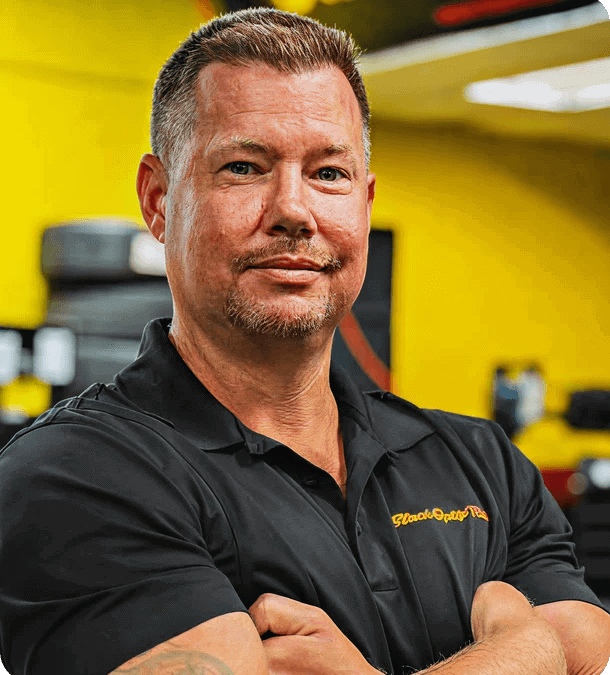 Self-confident man in black uniform with arms crossed in car repair shop, representing United Franchise Group automotive franchise opportunities and business growth.