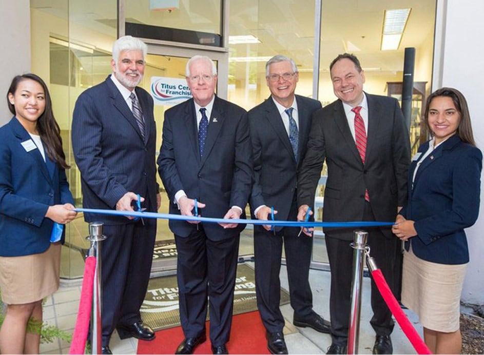 A group of diverse professionals participates in a ribbon-cutting ceremony outside a franchising business office with the United Franchise Group logo.